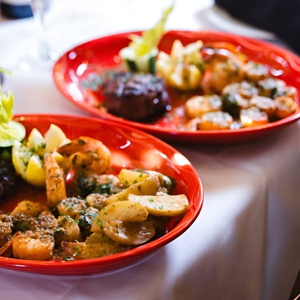 Two red plates of Bella Italia Ristorante's shrimp, potatoes, and steak on a white tablecloth.
