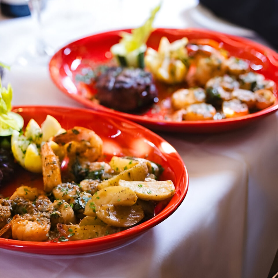 Two red plates of Bella Italia Ristorante's shrimp, potatoes, and steak on a white tablecloth.